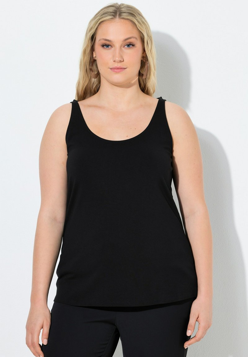 Blondynka z długimi włosami, nosząca kolczyki w kształcie kół i czarną, bezrękawnikową bluzkę, stojąca na jednolitym białym tle.