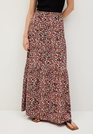 Femme portant une longue jupe fluide à fleurs roses, blanches et oranges, associée à un haut noir sans manches et des sandales.