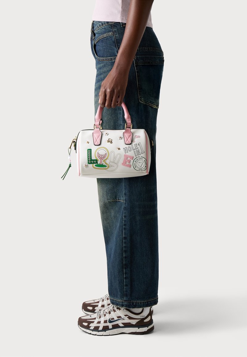 Personne portant un jean et des baskets tient un sac à main blanc avec des poignées roses et des écussons colorés sur le thème du golf.