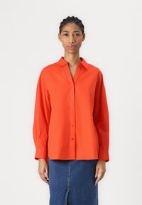 Camicia rossa con bottoni e colletto, maniche lunghe e vestibilità relaxed, abbinata a una gonna di jeans. Presenta un tessuto liscio e testurizzato.