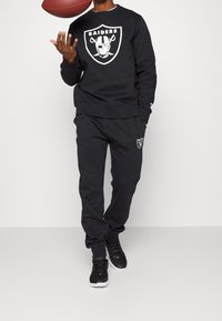 Felpa nera e pantaloni con il logo dei Raiders. Il tessuto ha una consistenza liscia. Sneakers nere con dettagli bianchi completano l'outfit.