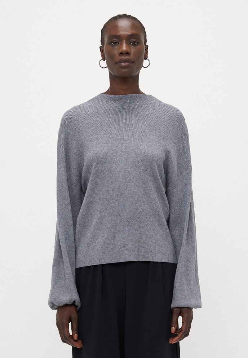 Grauer Strickpullover mit lockerer Passform, rundem Halsausschnitt und Ballonärmeln. Strukturierter Stoff, kombiniert mit schwarzen Hosen, minimalistische Details.