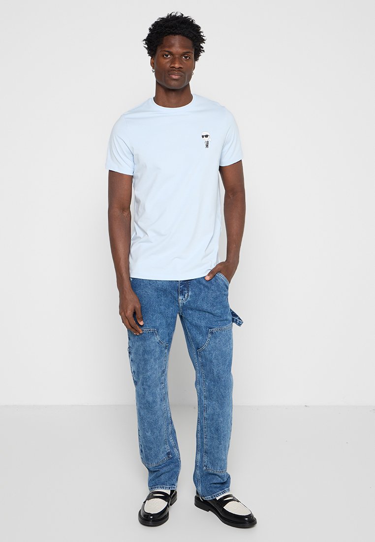Hombre con camiseta azul claro con pequeño gráfico, vaqueros azules con lazos laterales y zapatos blanco y negro, de pie contra un fondo blanco.
