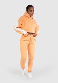 Kraftigt orange sweatshirt med hvide detaljer og joggebukser i matchende farve. Har hætte og afslappet pasform. Hvide sneakers.