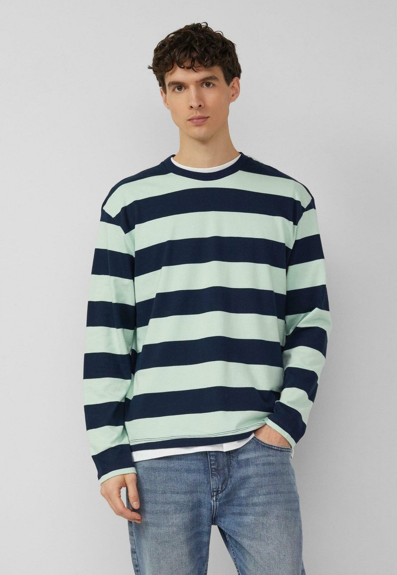 Nooruk kõhredaste lühikeste juustega, kes kannab pikkade varrukatega mintrohelist ja mereväe sinist triibulist särki ning helesiniseid teksapükse, seisab ilma taustata.