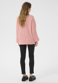 Übergroßer rosa Pullover aus weichem Strickstoff, mit weiter Ausschnittform und langen Ärmeln, kombiniert mit schmalen schwarzen Hosen und schwarzen Schuhen.