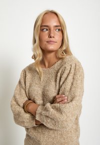 Bež, pahuljasti pleteni pulover s okruglim izrezom i napsuhanim rukavima. Tekstura izgleda mekana i ugodna, pogodna za hladnije vrijeme.