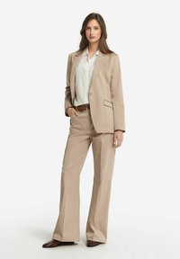 Beige skräddarsydd kostym med en enfärgad kavaj med slag och matchande utsvängda byxor. Vit blus och brun bälte ingår.