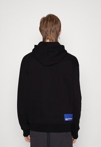 Sudadera negra con acabado texturizado, que presenta una gran capucha, mangas largas y un parche azul con el logo en el dobladillo.