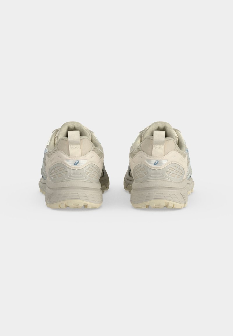 Par de zapatillas para correr por senderos de color beige, vistas desde atrás con suelas texturizadas visibles y lengüetas de tirador sobre un fondo liso claro.