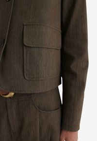 Brauner cropped Blazer mit zwei vorderen Taschen, texturiertem Stoff und sichtbaren Nähten. Passende braune Hose mit dezenten Taschendetails.