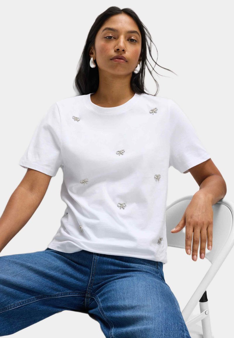 Bílý bavlněný tričko s dekoracemi v podobě stříbrných včel, kulatý výstřih, krátké rukávy, spárované s modrými džíny a bílou židlí.