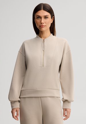 Femme aux cheveux bruns raides portant un sweat à capuche beige zippé et un pantalon assorti sur un fond clair uni.