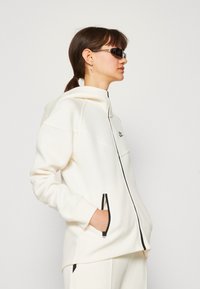 Baltos gobtuvinės zip-up striukės su šoninėmis kišenėmis, kuriomis puikuojasi juodi užtrauktuko akcentai ir logotipas kairėje krūtinės pusėje. Pagaminta iš minkštos, tamprios medžiagos.