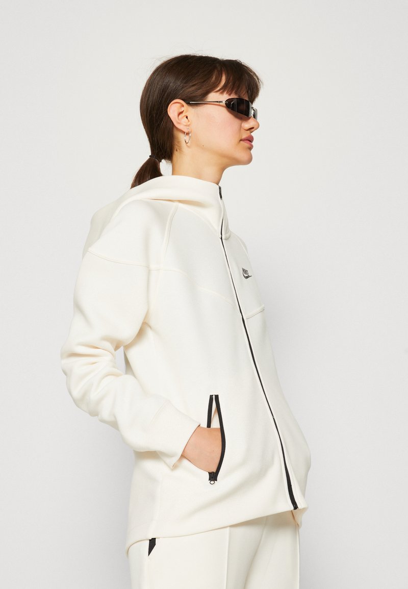 Chaqueta con capucha blanca, con cierre de cremallera y bolsillos laterales, que presenta detalles en negro en la cremallera y un logo en el lado izquierdo del pecho. Fabricada con un material suave y elástico.
