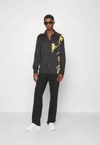 Zwarte langeklevende shirt met een gele sierlijke opdruk, gecombineerd met zwarte broek en witte sneakers. Het model draagt elegante zwarte zonnebril.