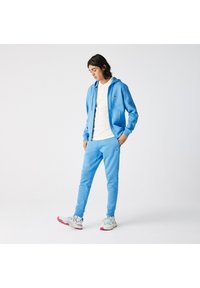 Lichtblauwe sweatshirt en bijpassende broek van zachte stof, met een capuchon, zijzakken en een embleem op de borst. Sneakers met veelkleurige accenten.