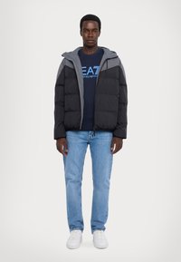 Giacca piumino nera e grigia con cappuccio, chiusura con zip e design trapuntato; indossata sopra una maglietta navy e jeans azzurri chiari con sneakers bianche.