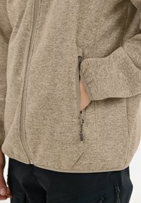 Beige texturerad jacka med dragkedja och sidofickor. Har ett subtilt mönster och kontrasterande kant längs dragkedjan.