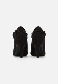 Scarpe con tacco alto in suede nero, caratterizzate da una forma curva, taglio alla caviglia e un accento decorativo a forma di fiocco sul lato. Superficie texturizzata con tacchi lisci.