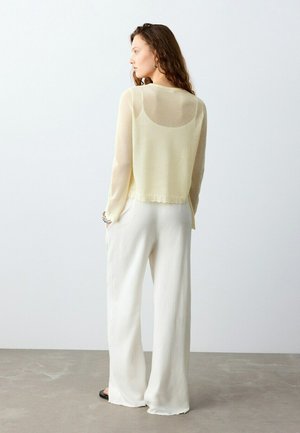 Donna con capelli lunghi che indossa una maglia beige trasparente a maniche lunghe e pantaloni bianchi larghi, in piedi con le mani nelle tasche, rivolta verso sinistra.