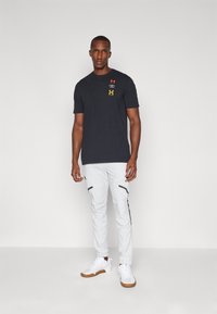 Crna majica kratkih rukava s tri otiska logotipa; kombinirana s uskim bijelim hlačama koje imaju crne zatvarače i bijelim tenisicama.