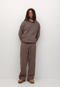 Bruine katoenen hoodie met oversized ontwerp, kangoeroezak, bijpassende relaxed-fit broek en beige schoeisel, tegen een neutrale achtergrond.