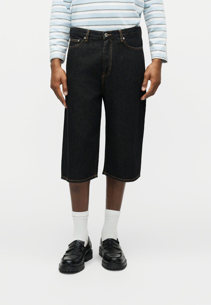 Fekete térdig érő farmernadrágot, fehér zoknit, fekete mokaszint és hosszú ujjú csíkos inget viselő személy áll egyszerű háttér előtt.