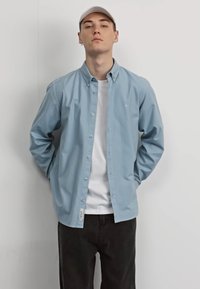 Chemise boutonnée bleu clair à manches longues, avec un col, et des boutons blancs, superposée sur un t-shirt blanc, associée à un jean noir.