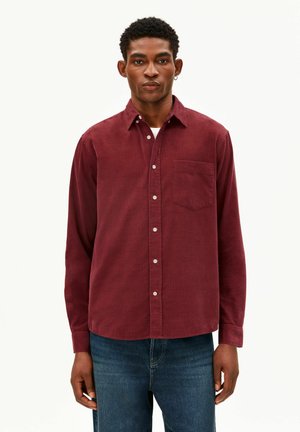 Uomo con una camicia di velluto a coste bordeaux con taschino sul petto e jeans blu, in piedi davanti a uno sfondo bianco semplice.