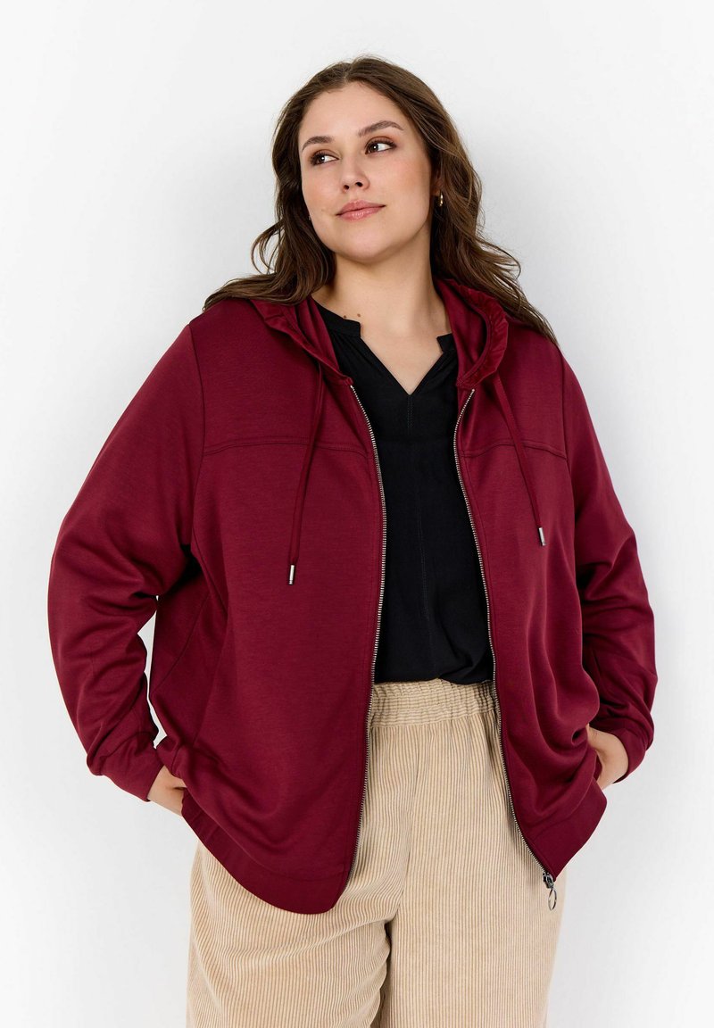Sudadera burdeos con cremallera y capucha con cordones, combinada con una camiseta negra y pantalones de pana beige, con un corte relajado.