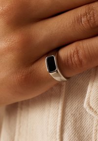 Zilveren ring met een rechthoekige zwarte steen, glad gepolijste band, gedragen op een vingertop tegen gestructureerde huid en stof.