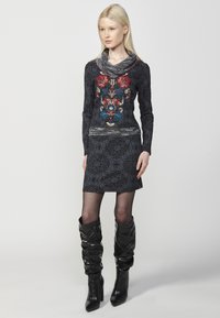 Schwarzes langärmliges Kleid mit floralem Stickerei in Rot und Blau, mit einem Wasserfall-Ausschnitt und einem filigranen Musterstoff. Kombiniert mit kniehohen schwarzen Stiefeln.