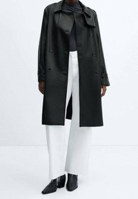 Schwarzer Leder-Trenchcoat mit doppelt geführtem Revers, vorderen Taschen und Knopfdetails, getragen über einem grauen Oberteil und weißen weit geschnittenen Hosen.