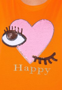 Orange t-shirt med et pinke paillet hjerte med et øje og vipper. Ordet "Happy" er broderet i guld under designet.