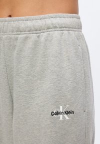 Κοντινή λήψη γκρι φόρμας με ελαστική μέση και το λογότυπο Calvin Klein κεντημένο κοντά στην τσέπη στο δεξί ισχίο.