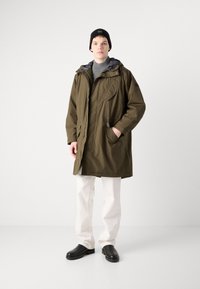 Parka impermeável verde oliva com capuz, fecho de correr e botão de pressão, bolso cargo frontal, combinado com um suéter cinza de gola alta e calças brancas.