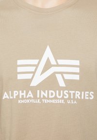 Tricou din bumbac bej cu un logo alb prezentând litere stilizate și linii orizontale. Textul spune "ALPHA INDUSTRIES KNOXVILLE, TENNESSEE, U.S.A."