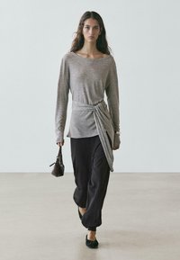 Szary top z długim rękawem z przodem zawiązanym w supeł, zestawiony z czarnymi spodniami o szerokich nogawkach. Modelka trzyma małą czarną torebkę, nosi płaskie buty.