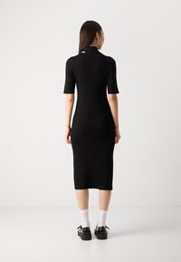 Abito midi nero in maglia con maniche corte, texture a costine e un sottile dettaglio del logo sul retro. Abbinato a scarpe nere e calze bianche.