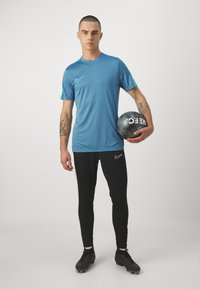 Sportovní oblečení zahrnující modré tričko Nike s krátkým rukávem a černé legíny, s fotbalovým míčem drženým v jedné ruce. Výrazné tetování na paži.