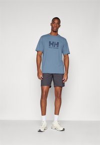 Gaiši zila īsām piedurknēm T-kreklu ar "HH" logotipu, apvienots ar tumšiem šortiem un bēšām sporta apavām. Redzami balti sporta zeķes.