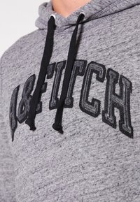 Primo piano di una felpa grigia con lacci neri e "A&FITCH" ricamato in grandi lettere nere sul petto.