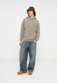 Haut à capuche en polaire gris clair avec une poche frontale, associé à un jean en denim bleu ample et des chaussures beiges. Une casquette noire complète le look.