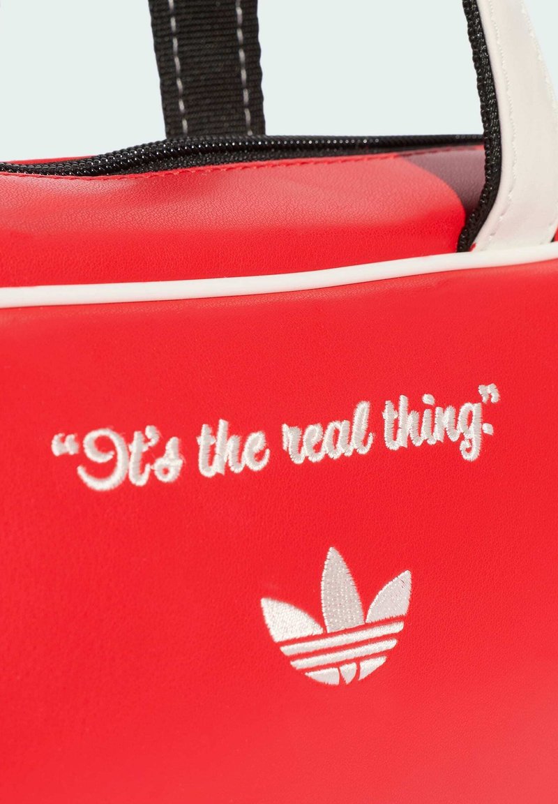 Red bag with black and white handles, embroidered text "It's the real thing" and a white Adidas trefoil logo below the text.