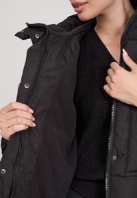 Giacca a vento nera con zip frontale, bottoni a pressione e morbida fodera interna. Indossata sopra un maglione di lana scuro, che mette in mostra dettagli testurizzati.