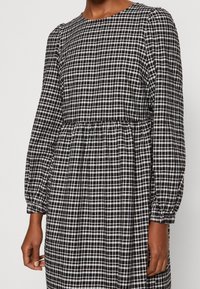 Svartvitt rutmönstrad klänning med långa, samlade ärmar och en figursydd midja. Tyget har en strukturerad, mjuk yta.