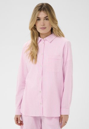Jeune femme aux longs cheveux blonds portant une chemise à rayures roses et blanches boutonnée et un pantalon assorti à rayures, sur un fond uni.