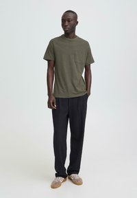 Mladić stoji u maslinasto zelenoj majici s džepom, crnim hlačama i bež tenisicama na jednostavnoj svijetloj pozadini.