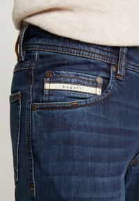 Jeans de mezclilla azul oscuro con una textura suave, que presentan una etiqueta blanca con "bugatti", costuras marrones y un detalle en el bolsillo frontal.